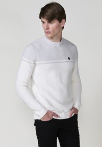 Witte gebreide trui met een gewoven patroon, voorzien van een ribbels ronde hals en manchetten, en een klein geborduurd logo op de borst.