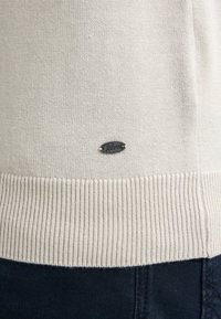 Suéter de punto beige con un dobladillo acanalado y una etiqueta de marca de metal negro con "Petrol" en la parte frontal. Textura suave y diseño casual.