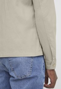Beige, long-sleeve shirt with a smooth texture and buttoned cuffs. Paired with light blue denim jeans featuring back pockets.
