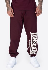 Bordeauxfarbene Jogginghose mit weißem "Lonsdale" Branding, mit Kordelzug, schmal zulaufendem Bein, aus weichem Baumwollmaterial. Getragen mit weißen Sneakers.