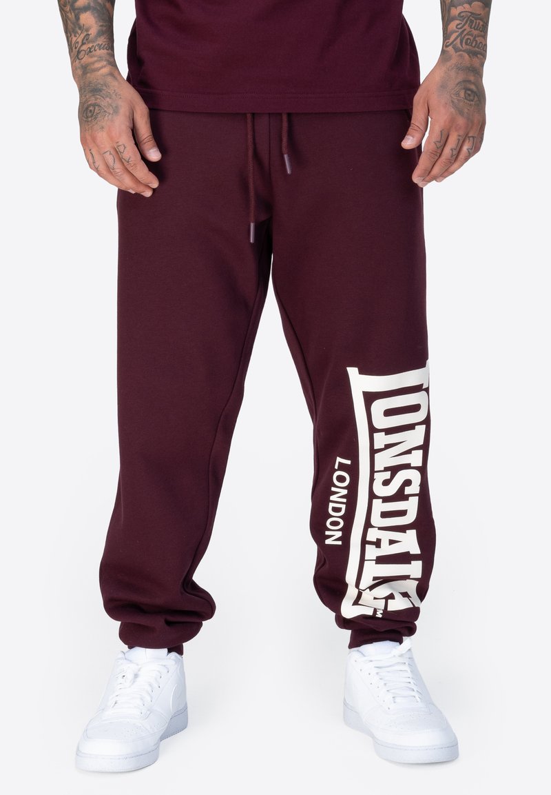 Bordeauxfarbene Jogginghose mit weißem "Lonsdale" Branding, mit Kordelzug, schmal zulaufendem Bein, aus weichem Baumwollmaterial. Getragen mit weißen Sneakers.