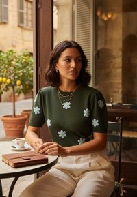 Kobieta z ciemnym bobkiem, ubrana w zielony sweter w białe kwiaty, siedzi przy stole z małą filiżanką kawy i brązowym notatnikiem w kawiarni.
