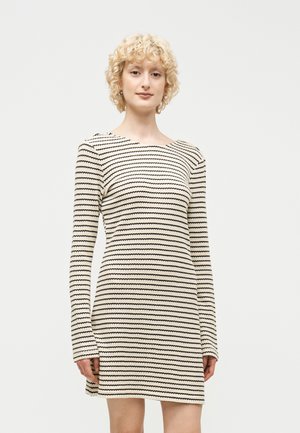 Lyshåret kvinde med krøllet hår iført en langærmet, cremefarvet og sort stribet strikkjole, stående mod en ensfarvet lys baggrund.