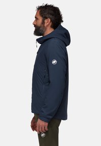 Marinblå isolerad jacka med huva, sidofickor med dragkedja och logotyp på ärmen, med en slät, matt yta.