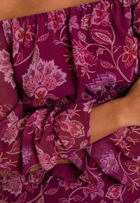 Off-the-shoulder blouse in diep magenta met bloemmotieven in roze en crème. Heeft geruite mouwen en een elastische taille voor een goede pasvorm.