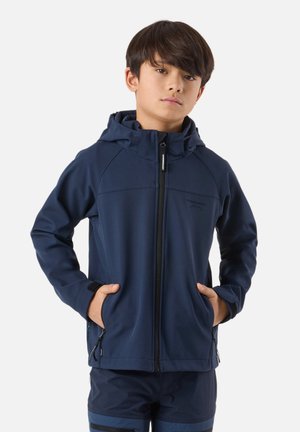 Ragazzo con giacca con cappuccio blu navy e pantaloni abbinati, in piedi con le mani nelle tasche davanti a uno sfondo bianco semplice.