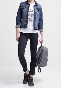 Chaqueta de mezclilla en azul oscuro, camiseta gráfica blanca, jeans ajustados oscuros, zapatillas negras, sosteniendo una mochila gris con textura suave.