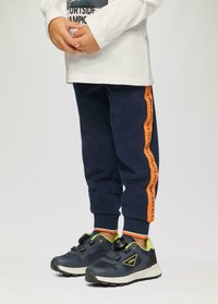 Marineblaue Jogginghosen mit orangefarbenem Seitenstreifen, der Text enthält. Kombiniert mit dunklen Sneakern mit grünen Akzenten und einem schwarzen Schnallenverschluss.