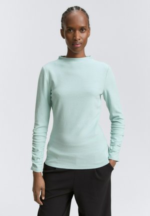 Femme avec des cheveux tressés portant un haut à manches longues bleu clair et un pantalon noir, mains dans les poches, se tenant devant un fond uni.