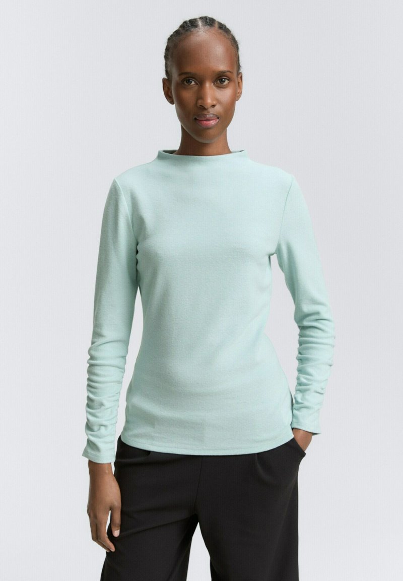 Kobieta z warkoczami, ubrana w jasnoniebieską bluzkę z długim rękawem i czarne spodnie, z rękami w kieszeniach, stojąca na tle jednolitym.