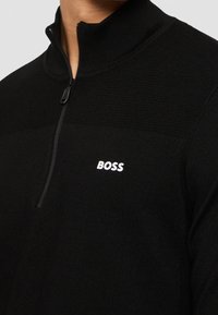 Black half-zip pullover made of textured fabric; features a high collar and white "BOSS" logo on the chest. Smooth, fitted design.