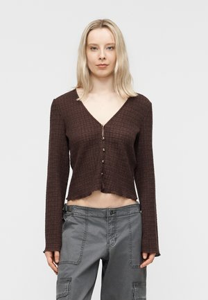 Donna bionda che indossa un top corto marrone testurizzato a maniche lunghe con dettagli a bottoni e pantaloni cargo grigi, in piedi davanti a uno sfondo uniforme.