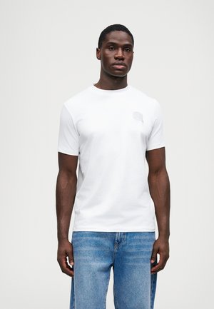 Weißes T-Shirt mit kurzen Ärmeln aus einem weichen Stoff, das auf der linken Brust mit einem dezenten Logo-Detail versehen ist, kombiniert mit hellblauen Jeans.