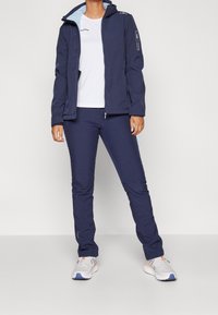 Veste imperméable marine avec capuche, associée à un pantalon marine. Les deux ont une texture lisse et des poches zippées. T-shirt blanc en dessous.