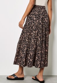 Falda maxi con estampado floral en tonos terrosos de topo y negro, con volantes y una cintura elástica. Combinada con sandalias negras tipo slide.