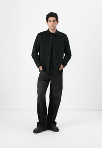 Dark green corduroy shirt with a button-up front, single chest pocket, paired with black wide-leg jeans and black shoes.