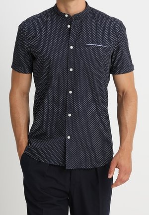 Mann trägt ein dunkelblaues kurzärmeliges Hemd mit kleinen weißen geometrischen Mustern und schwarze Hose, steht mit einer Hand in der Hosentasche.