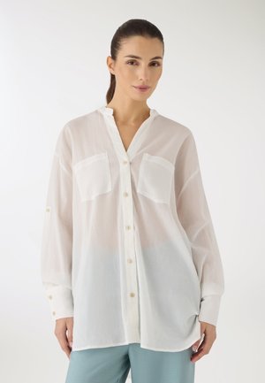 Donna con capelli scuri, indossa una camicia bianca trasparente con bottoni e pantaloni azzurri, in piedi davanti a uno sfondo bianco semplice.