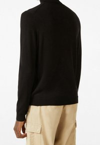 Svart stickad tröja med långa ärmar, ribbad halsringning och nederkant, kroppsnära passform, slät textur, sedd bakifrån. Kombinerad med beige cargobyxor.