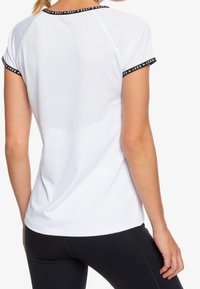 Mujer vestida con una camiseta atlética blanca de manga corta con ribetes negros y mallas negras, vista desde atrás contra un fondo simple.