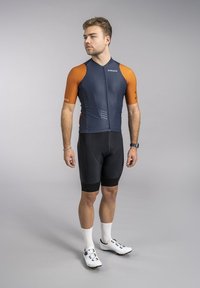 Cycling jersey in dark blue with orange sleeves, short front zipper, paired with black padded shorts and white cycling shoes.