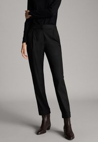 Schwarze maßgeschneiderte Hose mit geradem Bein, ab genähter Vorderseite und umgeschlagenem Saum, gefertigt aus einem glatten Stoff. Kombiniert mit einem enganliegenden schwarzen Oberteil und braunen Stiefeletten.