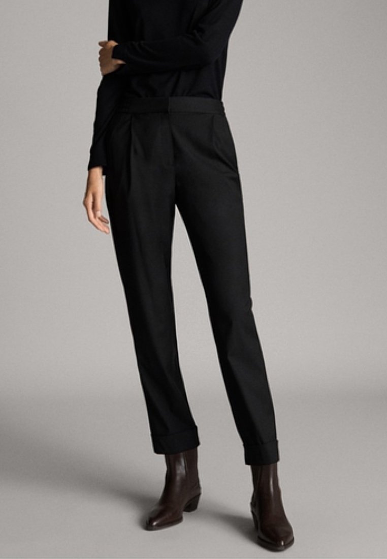 Schwarze maßgeschneiderte Hose mit geradem Bein, ab genähter Vorderseite und umgeschlagenem Saum, gefertigt aus einem glatten Stoff. Kombiniert mit einem enganliegenden schwarzen Oberteil und braunen Stiefeletten.