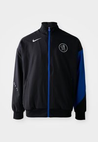 Crni sportski sako s plavim akcentima, s visokim ovratnikom, zatvaranjem na zip, bočnim džepovima i kružnim logotipom na prsima. Nike brendiranje.