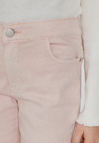 Světle růžové džínové kalhoty s přední kapsou, kovovým uzávěrem na cvaknutí a prošitými detaily podél švů a pásku.