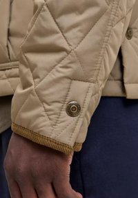 Manga de chaqueta beige acolchada con cierre de botón a presión metálico de marca y puño de pana marrón visible en una mano de piel oscura.
