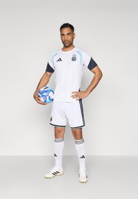 Λευκή ποδοσφαιρική φανέλα Adidas με ναυτικές μανίκες και ανοιχτό μπλε λεπτομέρειες, σε συνδυασμό με λευκά σορτς, μαύρες ρίγες και λευκά ποδοσφαιρικά παπούτσια, κρατώντας μια μπάλα ποδοσφαίρου.