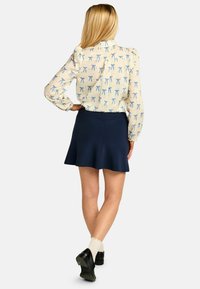 Majica s kremnimi dolgimi rokavi z modrim potiskom pentlje, kombinirana z razširjeno mornarsko modro krilo. Model nosi črne čevlje in kremne nogavice.