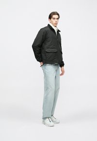 Casaco acolchoado preto com fecho de correr à frente, dois bolsos frontais, punhos elásticos e textura suave, usado sobre jeans azul claro e tênis.