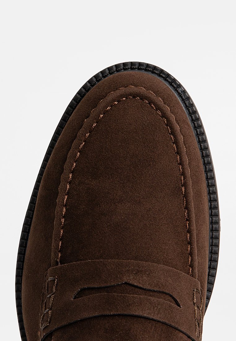 Mocasines de ante marrón con punta redonda, que presentan detalles de costura y una suela de cuero. La textura es suave, con acentos sutiles en la correa.
