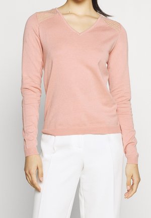 Kvinde iført lyserød V-hals sweater med blondedetaljer på skuldrene og lange ærmer, kombineret med hvide bukser.