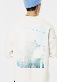 Persona che indossa una felpa bianca con una stampa sbiadita di un edificio urbano sulla schiena e un berretto di maglia azzurro chiaro, girata di spalle.