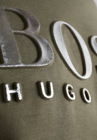 Primo piano del testo in rilievo argento "BOSS HUGO" su un tessuto verde oliva testurizzato.