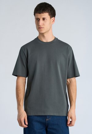 Ung mand med kort mørkt hår iført en ensfarvet mørkegrå kortærmet t-shirt og blå jeans, stående mod en lys baggrund.