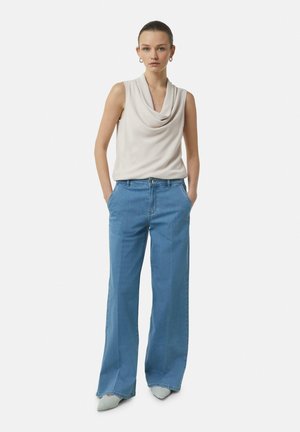 Top senza maniche di un bel beige chiaro con scollatura a cowl, abbinato a jeans larghi a vita alta di un blu chiaro. Le calzature presentano punte appuntite.