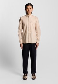 Licht beige overhemd met lange mouwen, knoopsluiting, een borstzak en een subtiel logo. Gepaard met donkere broeken en gestreepte sneakers.