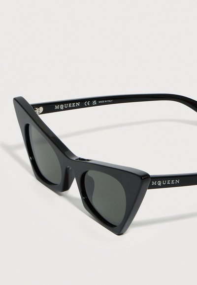 Μαύρα γωνιώδη γυαλιά ηλίου σε σχήμα γάτας με σκοτεινούς φακούς και το λογότυπο "McQueen" στα μπράτσα, απεικονιζόμενα σε κοντινή γωνία σε λευκό φόντο.
