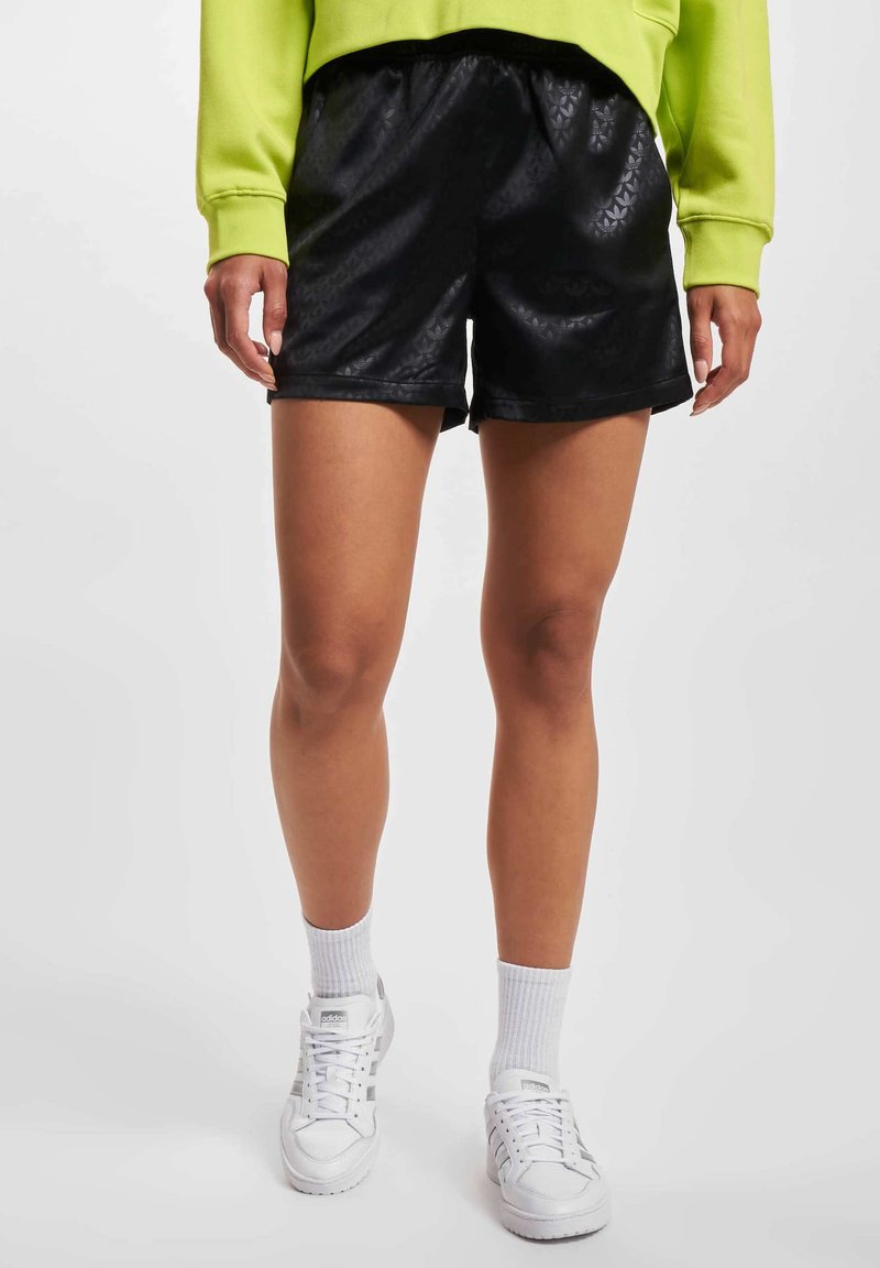 Personne portant un short noir à motifs, des baskets blanches avec des chaussettes blanches assorties, et un haut à manches longues vert vif.