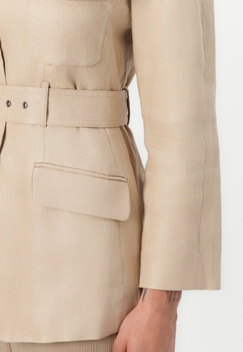 Beige Leinenjacke mit tailliertem Gürtel, ausgestattet mit einer aufgesetzten Tasche und langen gerade geschnittenen Ärmeln mit glatter Textur.