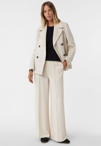 Doppelreihiger cremefarbener Wollblazer, schwarzes Oberteil und weite beige Hosen mit Seitennähten; das Modell steht mit einer Hand in der Tasche.