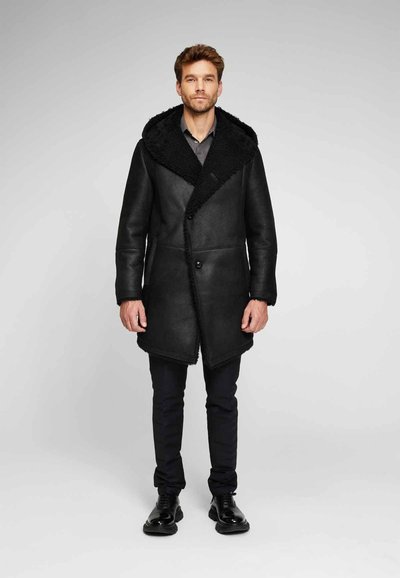 Cappotto di pelle nera con un ampio colletto in shearling testurizzato nero, chiusura a doppio petto e orlo asimmetrico. Indossato sopra pantaloni scuri.