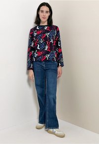 Donna in piedi contro un muro semplice, indossa un top lungo a maniche colorato con motivo, jeans svasati blu e sneakers bianche con dettagli blu.