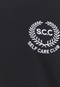 Tessuto nero con un logo ricamato bianco a forma di corona d'alloro con il testo "S.C.C" e "SELF CARE CLUB" al centro.