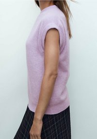 Pull à manches courtes violet clair en matière douce, avec un col rond et des accents côtelés, associé à une jupe noire à carreaux.