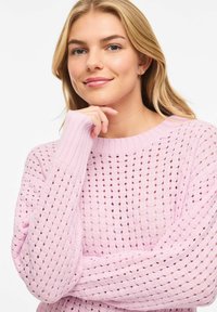 Jeune femme aux cheveux blonds portant un pull en tricot rose clair, reposant son menton sur sa main avec un léger sourire, fond blanc.