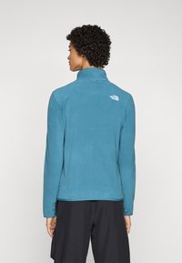 Lichtblauwe fleecejack met een hoge kraag en lange mouwen. Heeft een klein wit logo op de bovenrug. Zachte textuur, casual design.